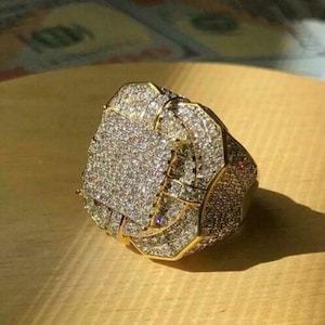 May include: A large, gold-toned ring encrusted with numerous small, sparkling clear stones. The ring has a geometric design with a square center and a textured surface. The ring is set on a light brown wooden surface.