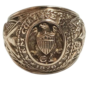 May include: A gold-colored ring with intricate detailing. The ring features an eagle emblem within a shield, surrounded by text that reads "COLLEGE OF THE TEXAS A&M 61 1876". The ring has a vintage or antique appearance.