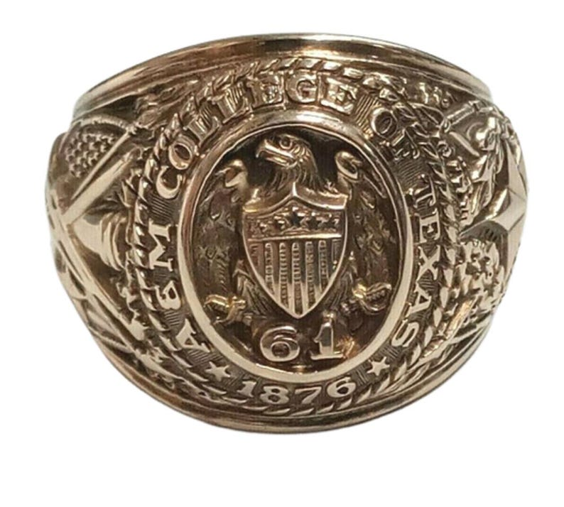 May include: A gold-colored ring with intricate detailing. The ring features an eagle emblem within a shield, surrounded by text that reads "COLLEGE OF THE TEXAS A&M 61 1876". The ring has a vintage or antique appearance.
