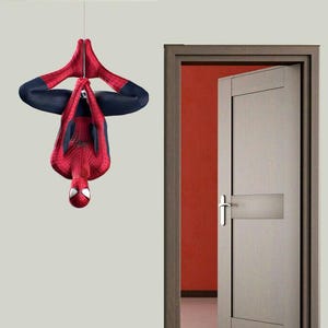 Può includere: Un adesivo da parete raffigurante un supereroe rosso e blu appeso a testa in giù tramite una linea sottile. Il personaggio indossa una tuta integrale con motivo a ragnatela, maniche lunghe e lenti oculari bianche, accanto a una porta in legno.