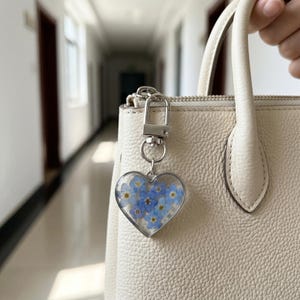 Puede incluir: Un llavero en forma de corazón con flores azules encapsuladas en resina transparente. El llavero está sujeto a un cierre plateado. El fondo incluye un bolso de mano color crema.