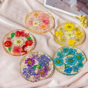 May include: Five round resin coasters with dried flowers embedded in clear resin. The coasters feature various flower arrangements in shades of pink, yellow, blue, purple, and red, with gold flakes. The coasters are displayed on a pink fabric surface.