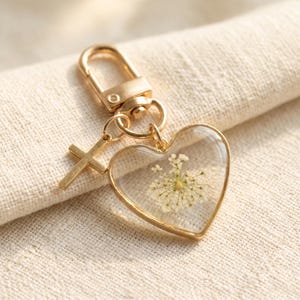 Pode incluir: Um chaveiro dourado com um pingente em forma de coração. O pingente tem um centro de resina transparente com flores brancas secas. Um pequeno pingente de cruz dourada pende ao lado do coração. O chaveiro está em um fundo de tecido bege.