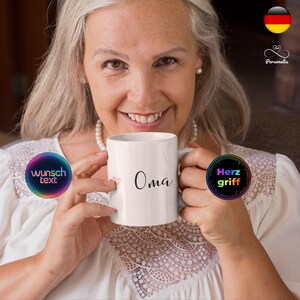 Könnte beinhalten: Eine weiße Keramiktasse mit dem Wort "Oma" in schwarzer Schrift. Die Tasse wird von einer Person mit grauem Haar gehalten, die ein weißes Oberteil und eine Perlenkette trägt. Die Tasse hat ein kleines Herzdesign neben dem Wort "Oma".