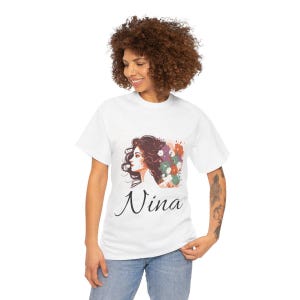 Pode incluir: Camiseta branca com uma ilustração em aquarela do perfil de uma mulher, cabelo escuro e flores coloridas. O nome "Nina" está escrito em itálico abaixo da imagem. A camiseta é usada por uma pessoa.