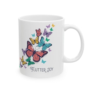 Może przedstawiać: Biały ceramiczny kubek z kolorowym motywem motyli. Motyle są w odcieniach różu, niebieskiego, żółtego, zielonego i fioletowego. Słowa "Flutter Joy" są napisane poniżej czcionką skryptową.