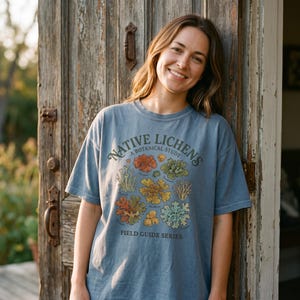 Op de afbeelding: Een leiblauw t-shirt met korte mouwen en een kleurrijke botanische afbeelding van diverse korstmossen. De tekst NATIVE LICHENS A BOTANICAL STUDY en FIELD GUIDE SERIES staat in donkergroene letters op de borst.