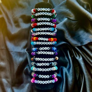 May include: A collection of colorful beaded bracelets with white letter beads spelling out various words. The bracelets feature a variety of colors, including red, blue, purple, and green. The words include "PRIMO", "SECONDO", "TERZO", and "CARDINA".