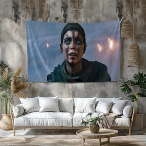 May include: A large tapestry featuring a digital painting of a person with blue eyes and dark markings on their face. The person is looking up, with a background of rain and torches. The tapestry is hanging on a concrete wall above a white couch.