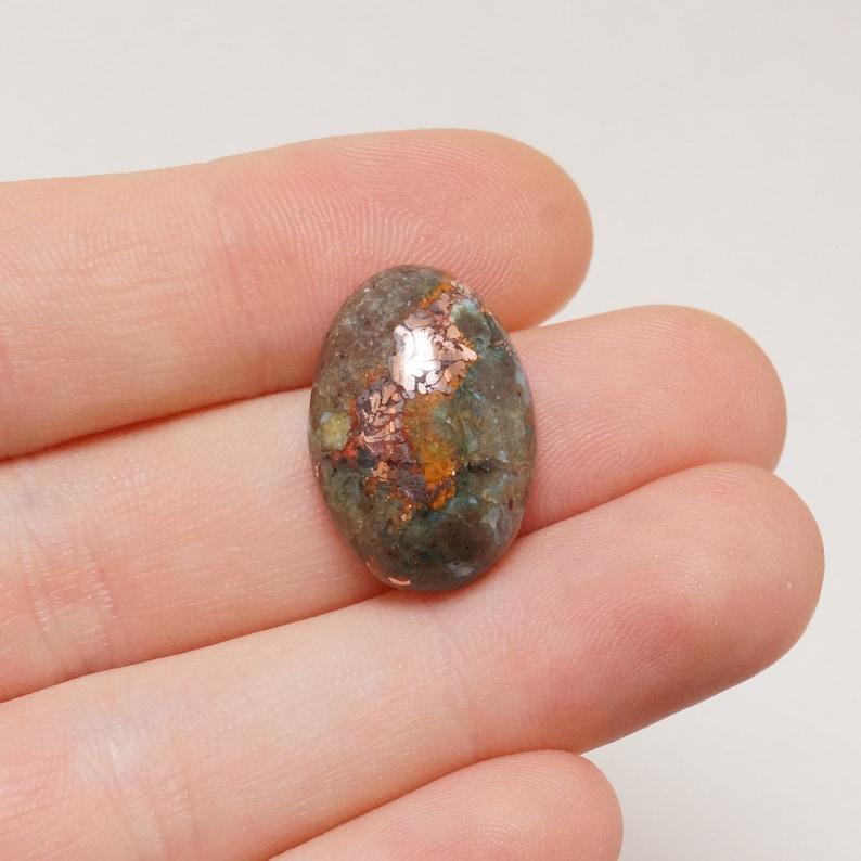 May include: Oval cabochon gemstone with a green, brown, and copper color pattern. The stone has a smooth, polished surface.