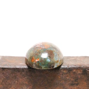 May include: A cabochon gemstone with a domed shape and a green, brown, and copper color pattern. The stone is sitting on a rusty metal surface.