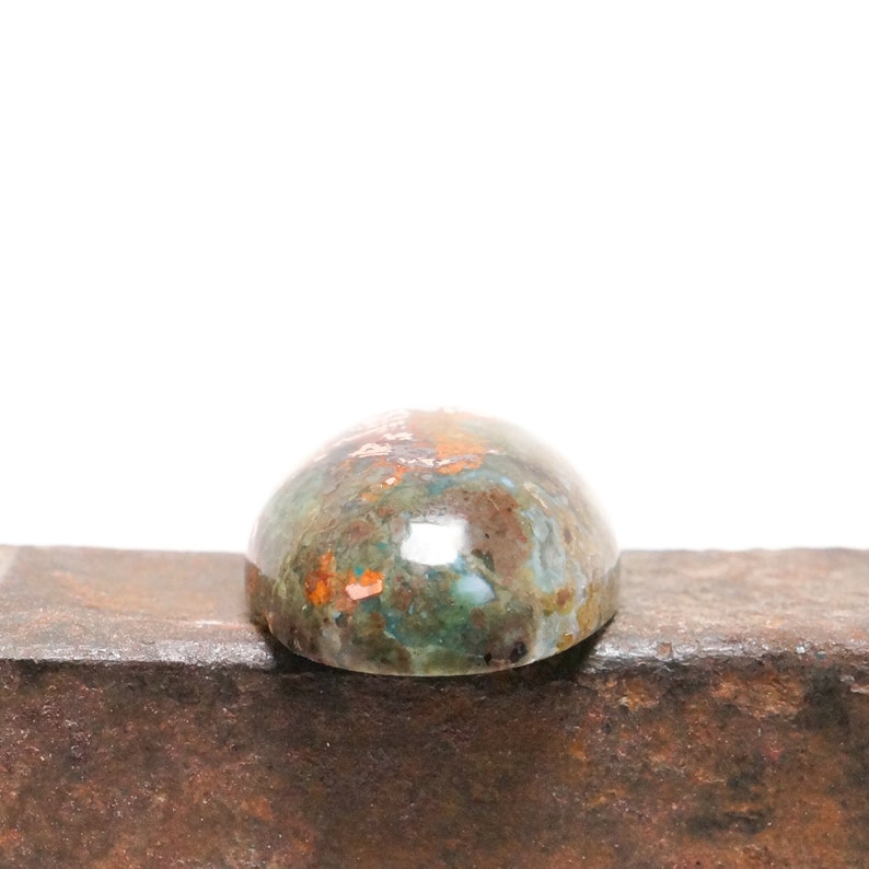 May include: A cabochon gemstone with a domed shape and a green, brown, and copper color pattern. The stone is sitting on a rusty metal surface.
