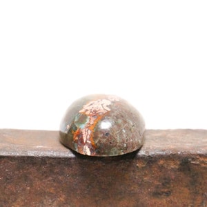 May include: A close-up of a green, brown, and copper-colored cabochon gemstone. The gemstone is oval-shaped and has a smooth, polished surface. It is sitting on a piece of metal.