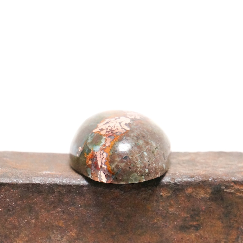 May include: A close-up of a green, brown, and copper-colored cabochon gemstone. The gemstone is oval-shaped and has a smooth, polished surface. It is sitting on a piece of metal.