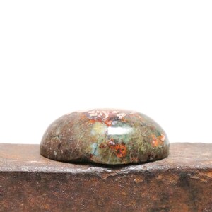 May include: A close-up of a green, brown, and orange cabochon gemstone with a smooth, polished surface. The gemstone is sitting on a piece of rough, rusty metal.