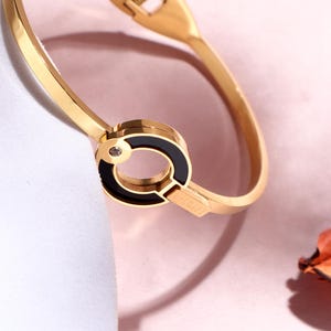 May include: Gold-tone bracelet with a black enamel accent and a small, clear gemstone. The bracelet features a hinged design with a clasp closure. The design includes the brand name. The jewelry is displayed on a pink and white surface.