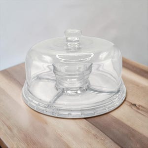 May include: A clear glass serving platter with a matching dome lid. The circular base features a central raised bowl section and a divided outer tray area. The lid has a small knob handle on top and a smooth, transparent finish throughout.