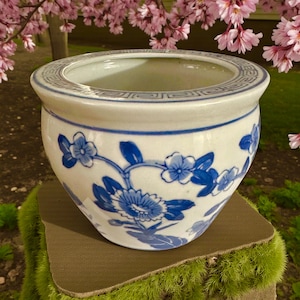 May include: A white ceramic planter with blue floral designs. The planter has a wide, round shape with a decorative border around the rim. The blue floral pattern includes flowers and leaves. The planter is sitting on a brown surface.