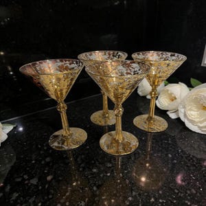 May include: A set of four martini glasses featuring a metallic gold mottled finish on the bowls and stems. Each glass has a wide, V-shaped bowl and a slender stem with a decorative knob detail, resting on a circular base with matching gold accents.