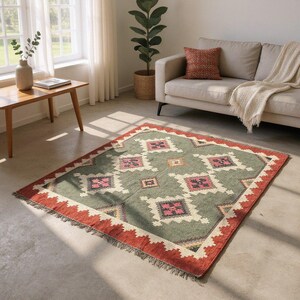 May include: A rectangular rug with a geometric pattern. The rug has a sage green field with diamond shapes in white, pink, and black. A red border with a zig-zag design frames the rug, and it has a fringed edge.