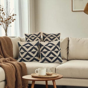 May include: Four decorative throw pillows with a geometric pattern in navy blue and beige are arranged on a beige sofa. A brown throw blanket and a small wooden table with a coffee cup and tray are in the foreground.