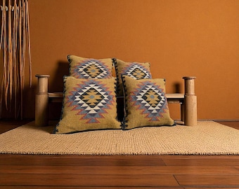 May include: Four decorative throw pillows with a geometric pattern in shades of gold, blue, gray, and black. The pillows are arranged on a small wooden bench, set on a woven rug, against a warm orange wall.