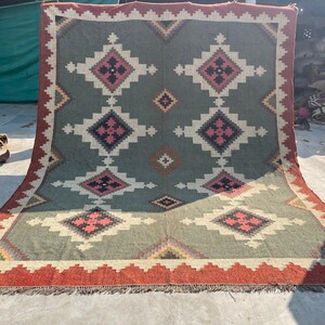 May include: A large, rectangular rug with a geometric pattern. The rug features a central design of repeating diamond shapes in shades of green, cream, and pink, framed by a red border with fringe. The rug is suitable for home decor.
