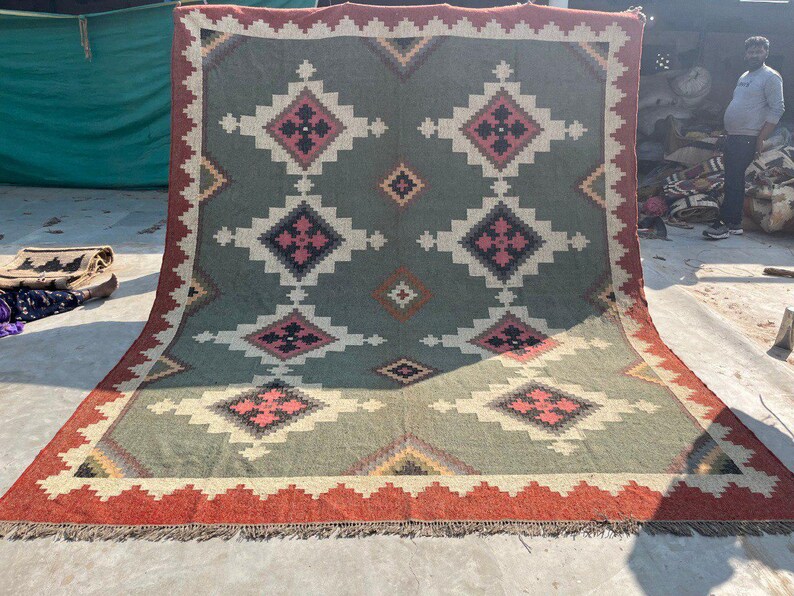 May include: A large, rectangular rug with a geometric pattern. The rug features a central design of repeating diamond shapes in shades of green, cream, and pink, framed by a red border with fringe. The rug is suitable for home decor.