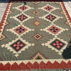 May include: A large, rectangular rug with a geometric pattern. The rug features a central field of muted green with repeating diamond shapes in pink, black, and white. The border is a deep red with a zig-zag design. The rug is suitable for a living room or bedroom.