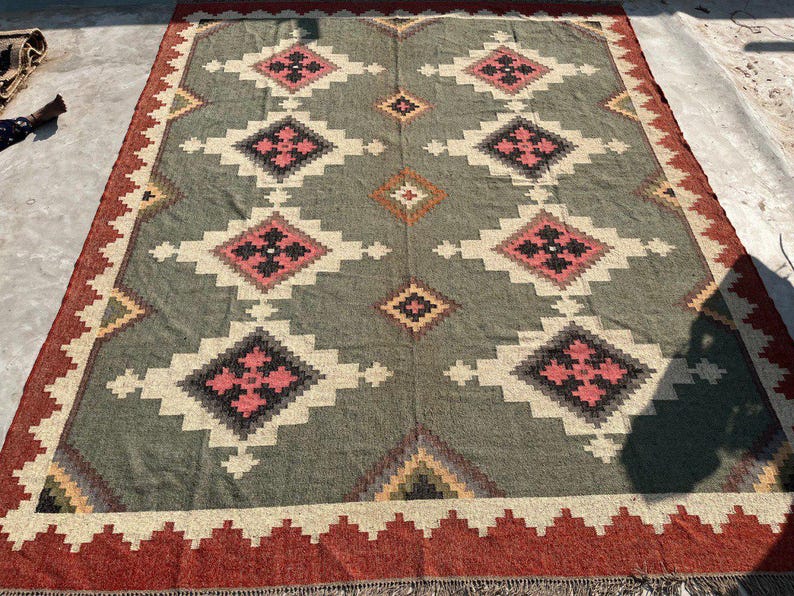 May include: A large, rectangular rug with a geometric pattern. The rug features a central field of muted green with repeating diamond shapes in pink, black, and white. The border is a deep red with a zig-zag design. The rug is suitable for a living room or bedroom.