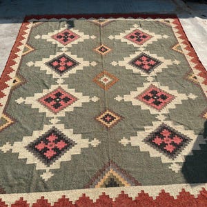 May include: A large, rectangular rug with a geometric pattern. The rug features a central field of grey with repeating diamond shapes in white, black, and pink. The border is a deep red with a zig-zag design. A decorative floor covering.