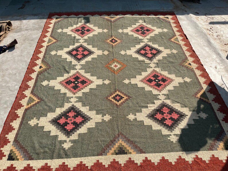 May include: A large, rectangular rug with a geometric pattern. The rug features a central field of grey with repeating diamond shapes in white, black, and pink. The border is a deep red with a zig-zag design. A decorative floor covering.