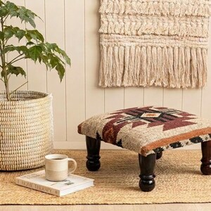 May include: A square footstool featuring a woven fabric top with a geometric pattern in shades of brown, cream, and black, supported by four dark brown, turned wooden legs. The stool is positioned near a woven basket containing a green plant.