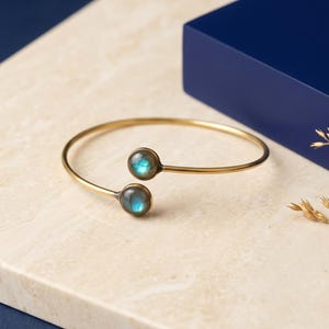 May include: A gold-toned open cuff bracelet featuring two round, cabochon-cut labradorite stones at each end. The stones display a blue iridescent flash, and the metal band is thin and circular, creating a simple, adjustable design.