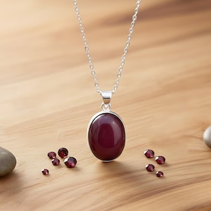 Könnte beinhalten: Eine silberne Halskette mit einem großen, ovalen, tiefroten Anhänger. Kleine, lose, rote Edelsteine sind um die Halskette auf einer Holzoberfläche verstreut. Die Halskette ist ein schlichtes, elegantes Design.