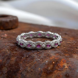 Puede incluir: Un brazalete plateado con piedras preciosas de rubí ovaladas y pequeños diamantes blancos. El brazalete tiene un diseño intrincado y detallado y está sobre una superficie oxidada y texturizada.