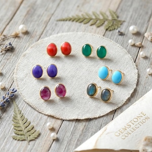 Puede incluir: Una colección de pendientes de botón con piedras preciosas en varios colores, incluyendo rojo, verde, morado, turquesa y rosa. Cada par tiene un engaste dorado. Los pendientes están dispuestos sobre una tela circular de color neutro, con una tarjeta que dice "GEMSTONE COLLECTION".