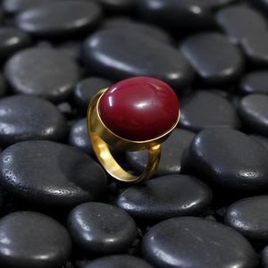 Op de afbeelding: Een gouden ring met een grote, ronde, dieprode steen. De band en zetting van de ring zijn helder goud van kleur, wat contrasteert met de donkerrode steen. De ring wordt getoond op een bed van gladde, zwarte stenen.