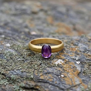 May include: A gold ring with a large, oval-cut purple amethyst gemstone. The ring has a simple, smooth band and is set against a textured, gray and green stone surface.