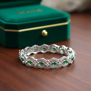 Op de afbeelding: Een zilveren armband met een herhalend patroon van ovale groene edelstenen en kleine witte stenen. De armband ligt op een houten oppervlak, met een groene fluwelen sieradendoos op de achtergrond.