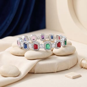 May include: A silver-toned bracelet featuring a series of rectangular-cut gemstones in various colours including red, green, blue, pink, and purple, alternating with small clear round stones, all set within a textured, beaded metal frame.