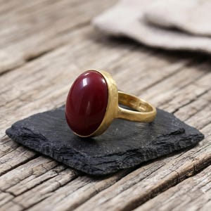 Op de afbeelding: Een gouden ring met een grote, gladde, ovaalvormige dieprode steen in een getextureerde metalen band. De ring heeft een gepolijste afwerking op de edelsteen en een matte, gehamerde uitstraling op de metalen zetting van het sieraad.