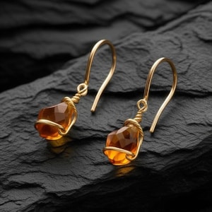 May include: A pair of gold drop earrings. Each earring has a faceted, amber-coloured gemstone wrapped in gold wire and suspended from a curved gold ear wire. The earrings are set against a dark, textured background.