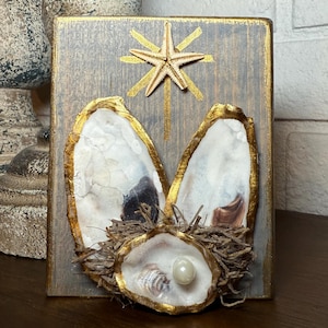Può includere: Un blocco di legno con un lavaggio grigio e finiture dorate presenta un design a stella marina e conchiglia di ostrica. Le conchiglie di ostrica sono disposte per assomigliare a orecchie di coniglio, con una perla al centro. Un pezzo decorativo per Pasqua o l'arredamento costiero.