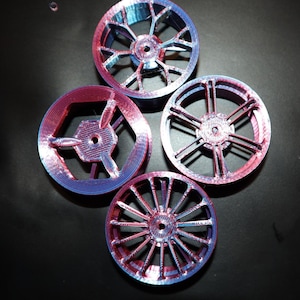 May include: Four 3D-printed wheels with a gradient of red and blue colors. Each wheel has a unique spoke design, with a central hub. The wheels are arranged on a dark surface, showcasing their intricate details.