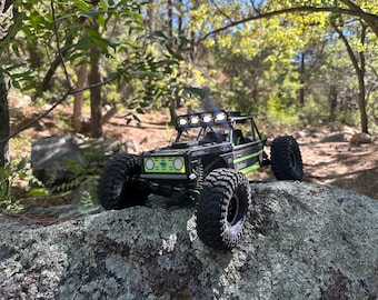 May include: A black and green Ford Bronco RC rock crawler with large, knobbly tyres and a light bar. The toy vehicle is positioned on a large rock in a natural outdoor setting, surrounded by trees and foliage.