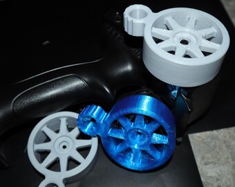 Puede incluir: Un transmisor RC negro con tres accesorios de rueda impresos en 3D en gris y azul. Las ruedas tienen un diseño de radios en forma de estrella. Los accesorios están diseñados para mejorar el agarre y el control del transmisor.
