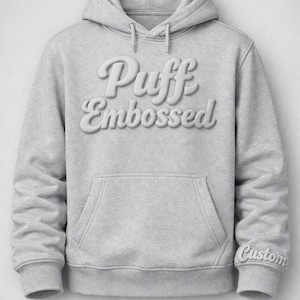 May include: A light gray hoodie with the words "Puff Embossed" in a puffy, raised font across the chest. The hoodie has a front pocket and a drawstring hood. The word "Custom" is embroidered on the cuff.