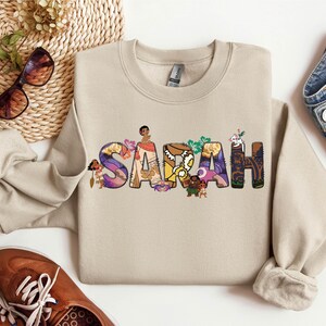 May include: A beige crewneck sweatshirt with the name "SARAH" printed across the front in colorful, stylized letters. Each letter is decorated with images of characters and floral designs. The sweatshirt is paired with blue jeans and brown shoes.