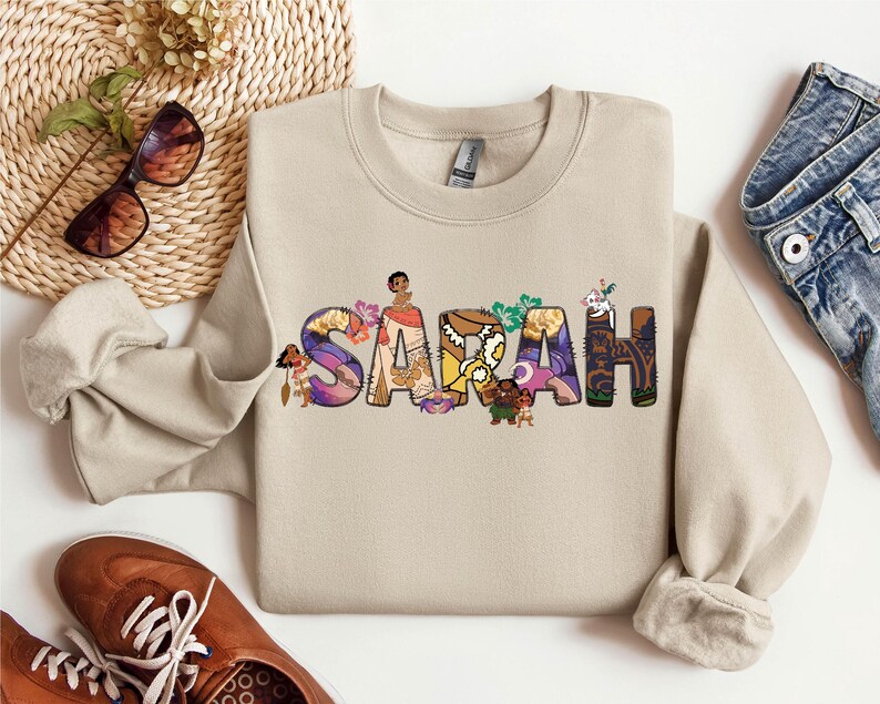 May include: A beige crewneck sweatshirt with the name "SARAH" printed across the front in colorful, stylized letters. Each letter is decorated with images of characters and floral designs. The sweatshirt is paired with blue jeans and brown shoes.